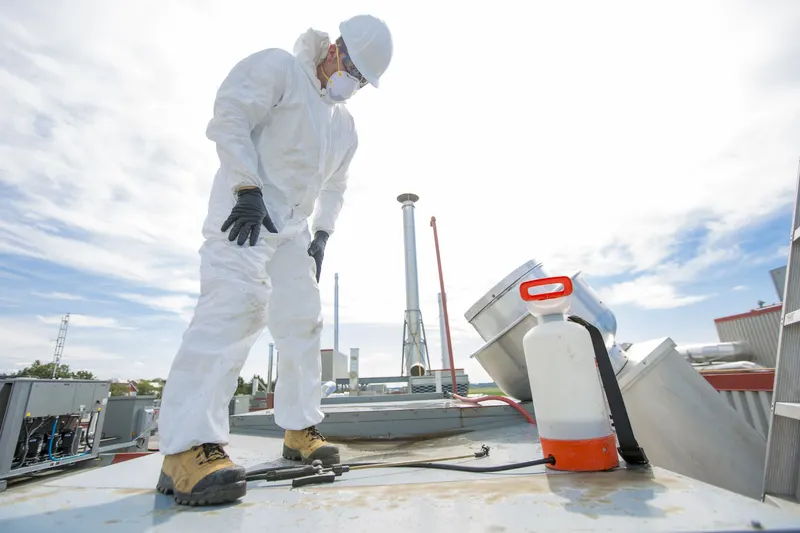 Technician in a hard hat standing on a flat roof beside an exhaust fan and a pump sprayer