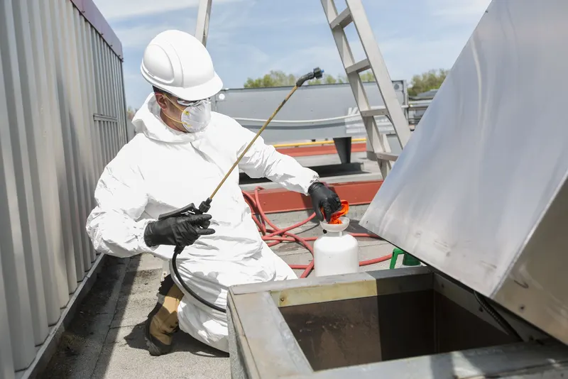 Technician kneeling on a roof spraying the base of a tipped-open exhaust fan with a pump sprayer