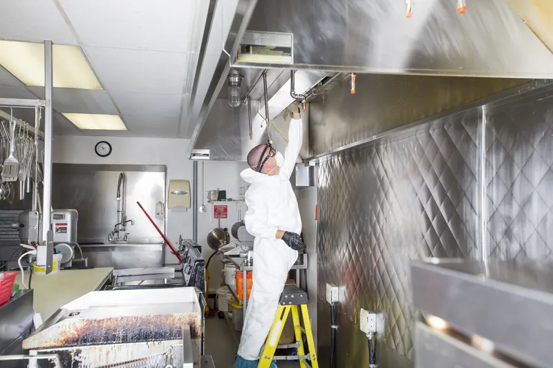 Technician on a yellow step ladder spraying the inside of a long stainless exhaust canopy above a commercial cooking line