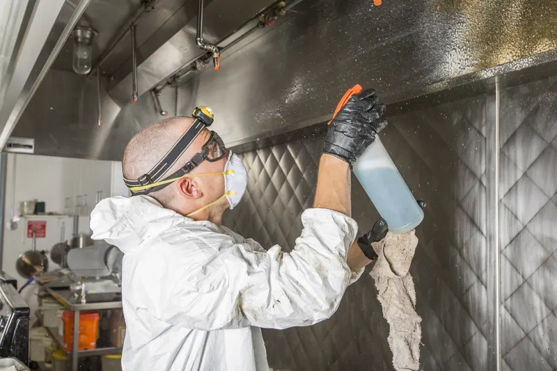 Technician in goggles and a mask spraying degreaser up into a stainless canopy while holding a cloth