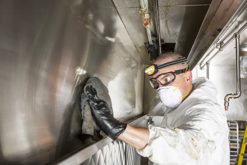 Technician in goggles and a mask wiping grease from the inside wall of an exhaust canopy
