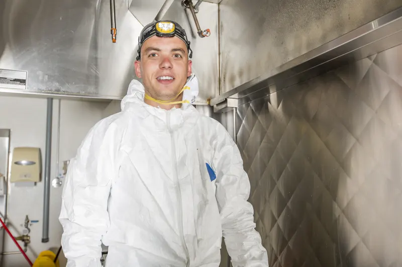 Hood cleaning technician in white coveralls and a headlamp standing under a stainless exhaust canopy