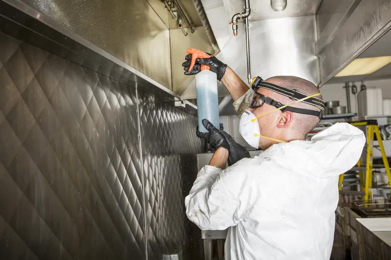 Technician spraying cleaner inside a canopy with a commercial kitchen and a step ladder behind
